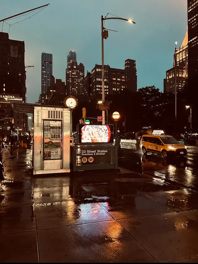 A wet New York street at dusk by the 23 Street subway station, with a yellow cab