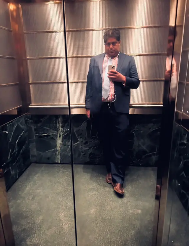 Kareem Abdo in a suit, taking a mirror photo in a marble and steel elevator